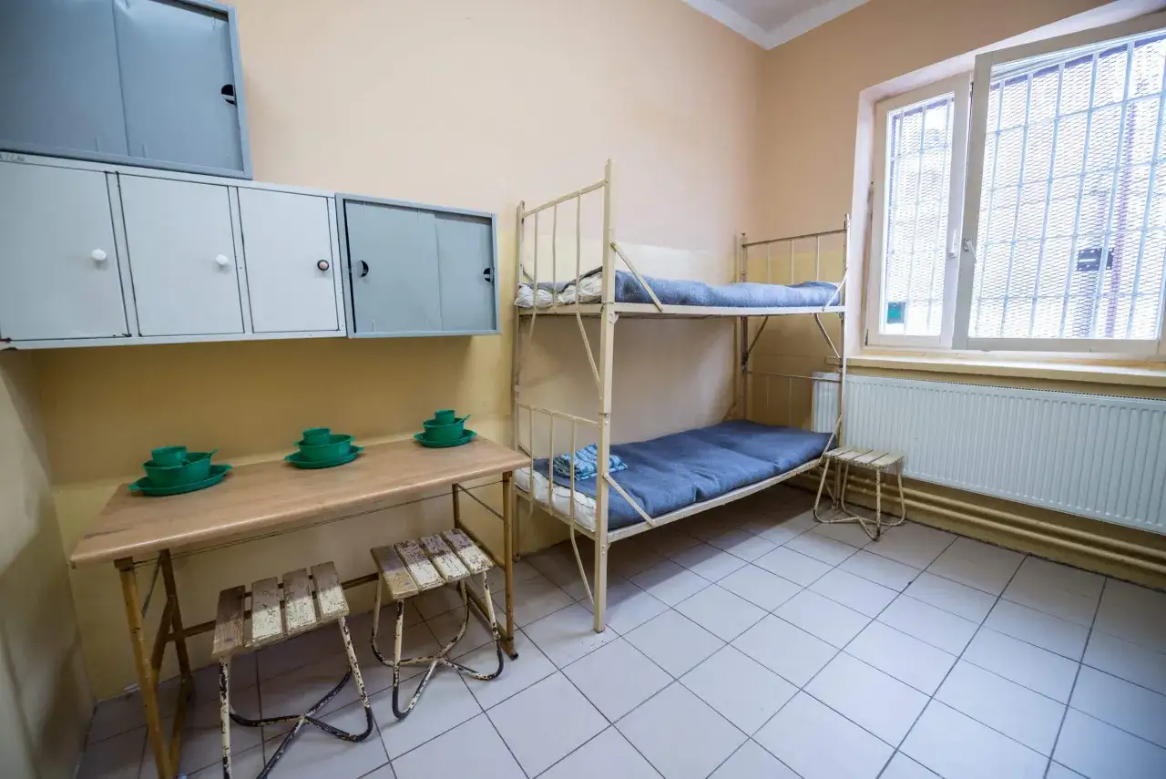 Cela wygląda jak cela w areszcie Montelupich. W pokoju są dwa łóżka piętrowe, stół z zielonymi naczyniami i szafki.