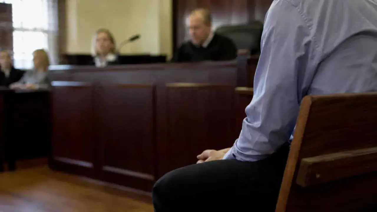 Osoba siedzi w ławie sądowej, czekając na rozprawę. W tle sędzia i ławnicy. To scena z sali sądowej, gdzie zapadają ważne decyzje, zgodne z art. 37a.