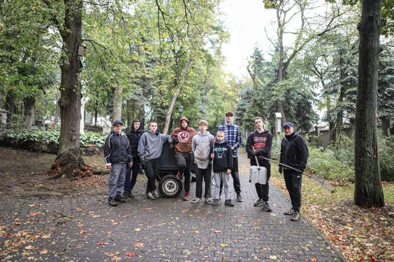 Grupa młodych ludzi, być może z ośrodka resocjalizacyjnego, pracuje na zewnątrz. W tle drzewa i jesienne liście.