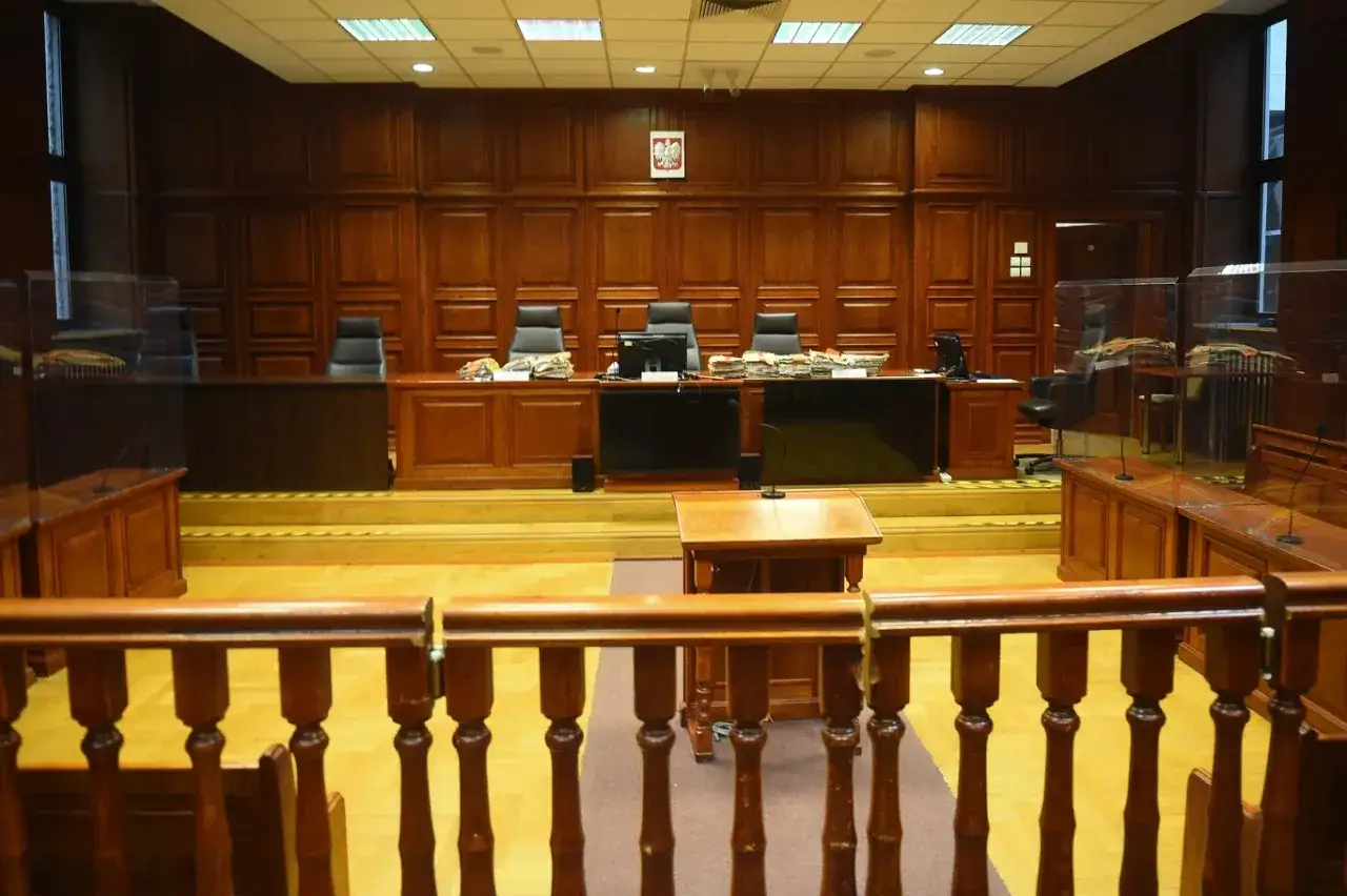 Pusta sala sądowa, gotowa na rozpatrzenie wniosku o zadośćuczynienie. Drewniane ławy, stoły z aktami i godło Polski.