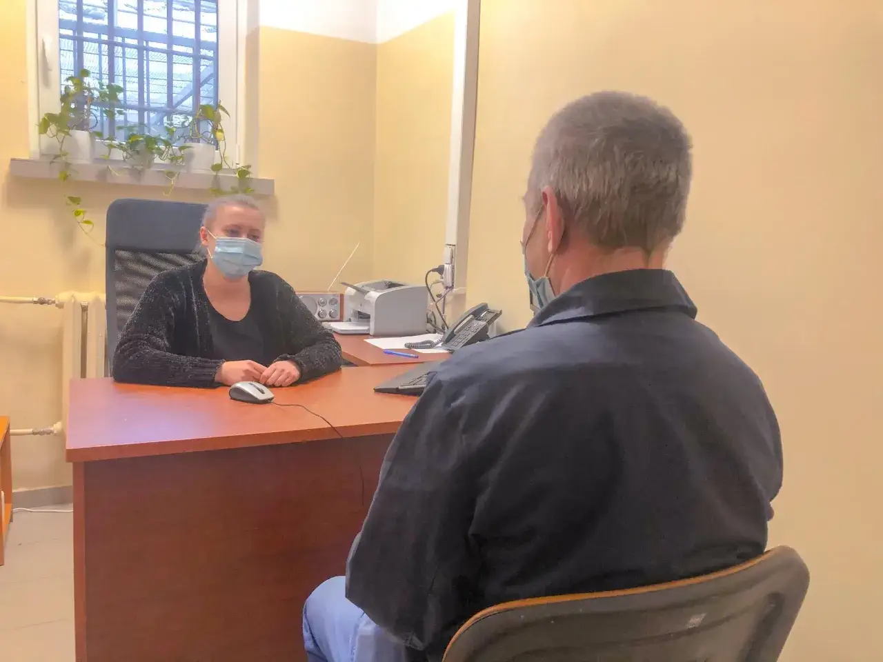 Psycholog więzienny w masce rozmawia z osadzonym. W tle okno z kratami i rośliny.