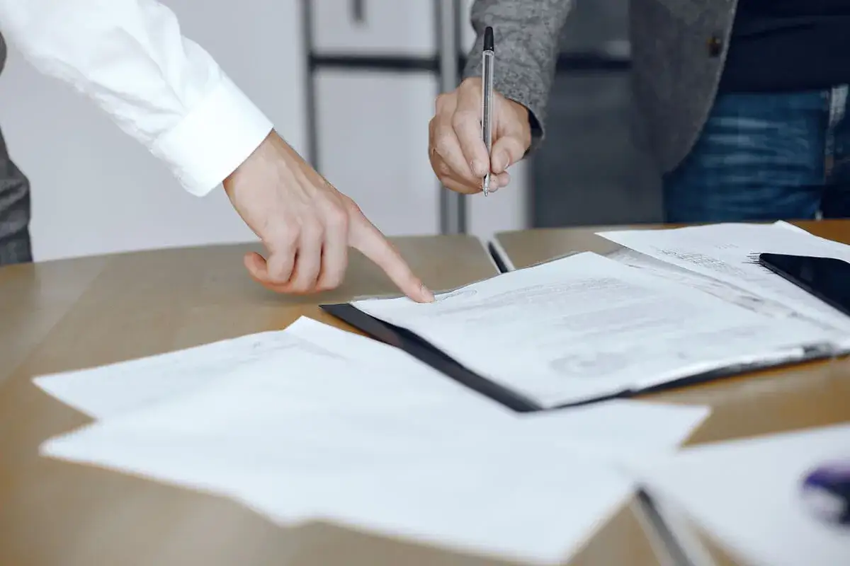 Dwie osoby przy biurku analizują dokumenty. Jedna wskazuje palcem na tekst, druga podpisuje. Może to być list z prokuratury, umowa lub ważny protokół.