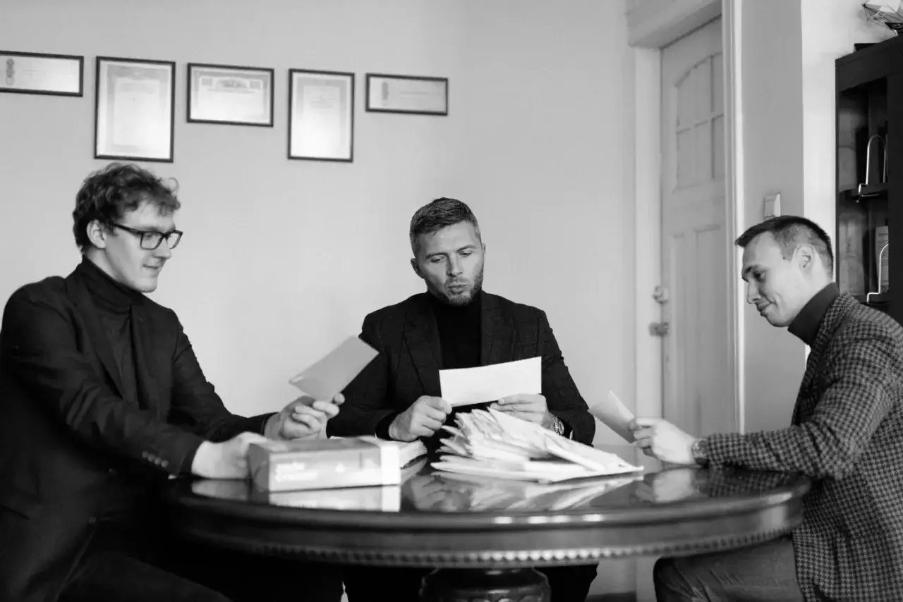 Trzech mężczyzn przy okrągłym stole analizuje dokumenty. Jeden z nich trzyma kopertę, zastanawiając się, co to znaczy 1 rok w zawieszeniu na 3 lata.
