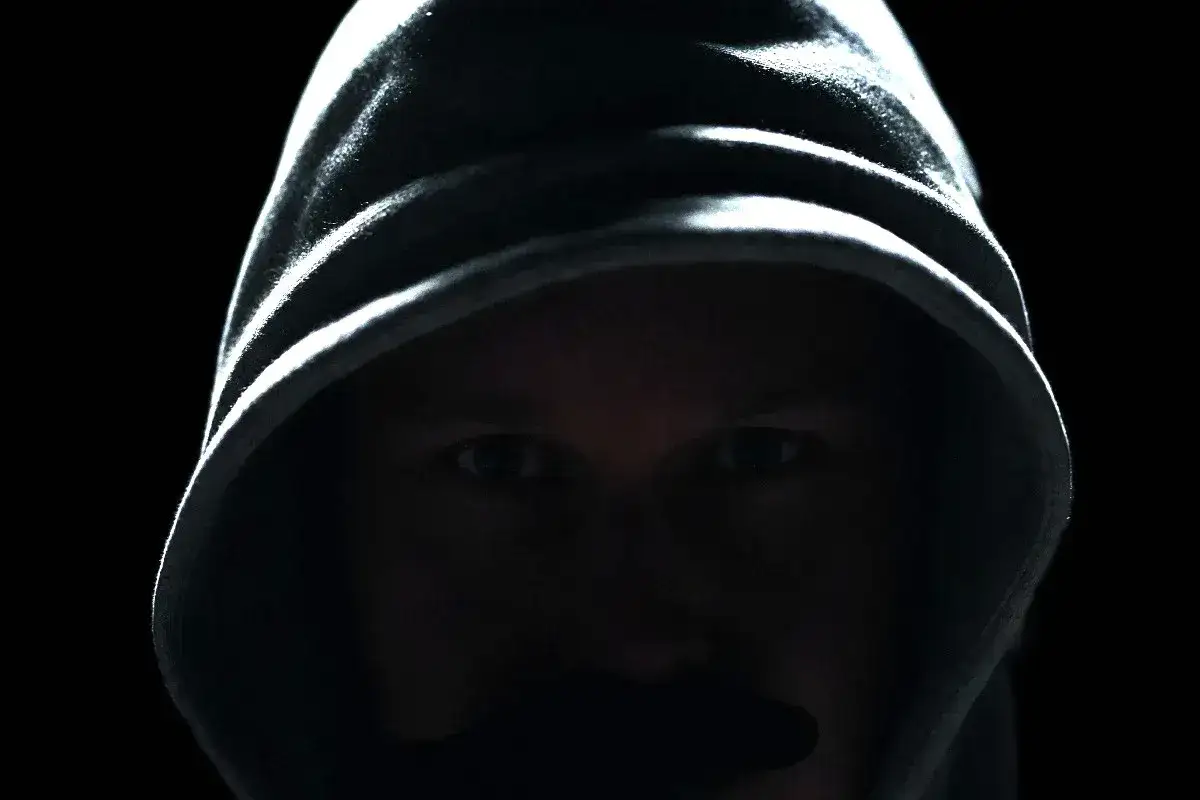 Osoba w kapturze, z ukrytym pod nim obliczem, czujnie obserwuje. Widać tylko oczy, sugerujące śledzenie kogoś.