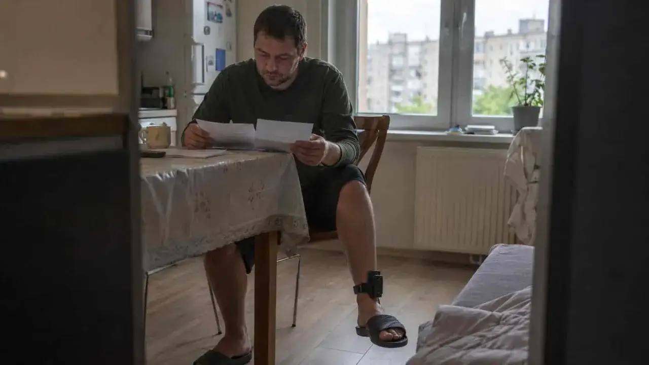 Mężczyzna z opaską dozoru elektronicznego na nodze przegląda dokumenty przy stole. Może to być związane z wynajmem mieszkania lub innymi formalnościami.