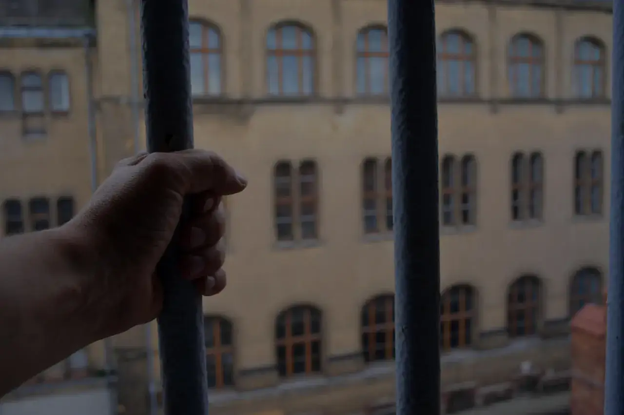 Dłoń zaciska się na kratach. Za nimi widać budynek z oknami, symbolizujący prawa i obowiązki skazanego.