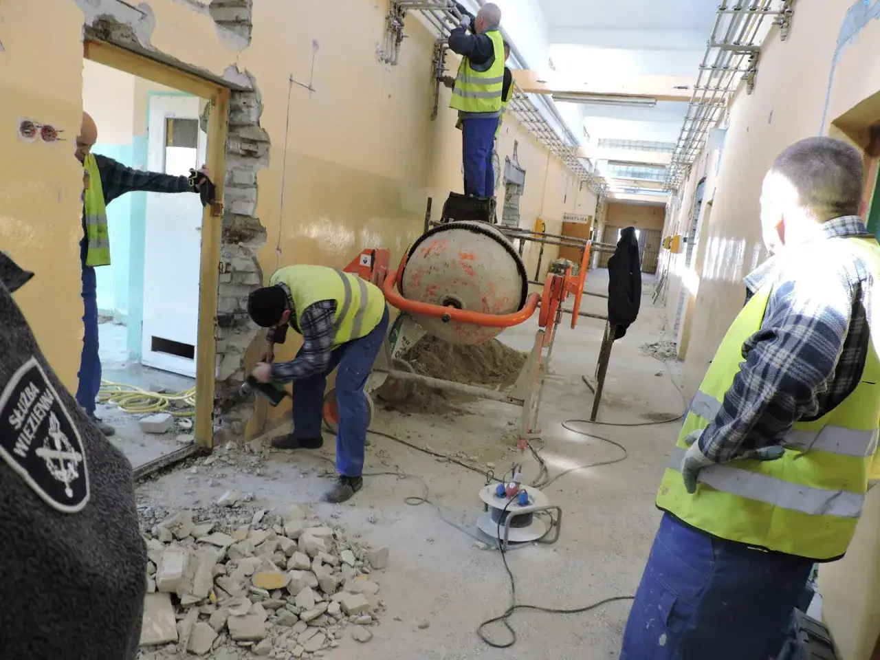 Praca w więzieniu: remont korytarza, gruz, betoniarka i pracownicy w kamizelkach odblaskowych.