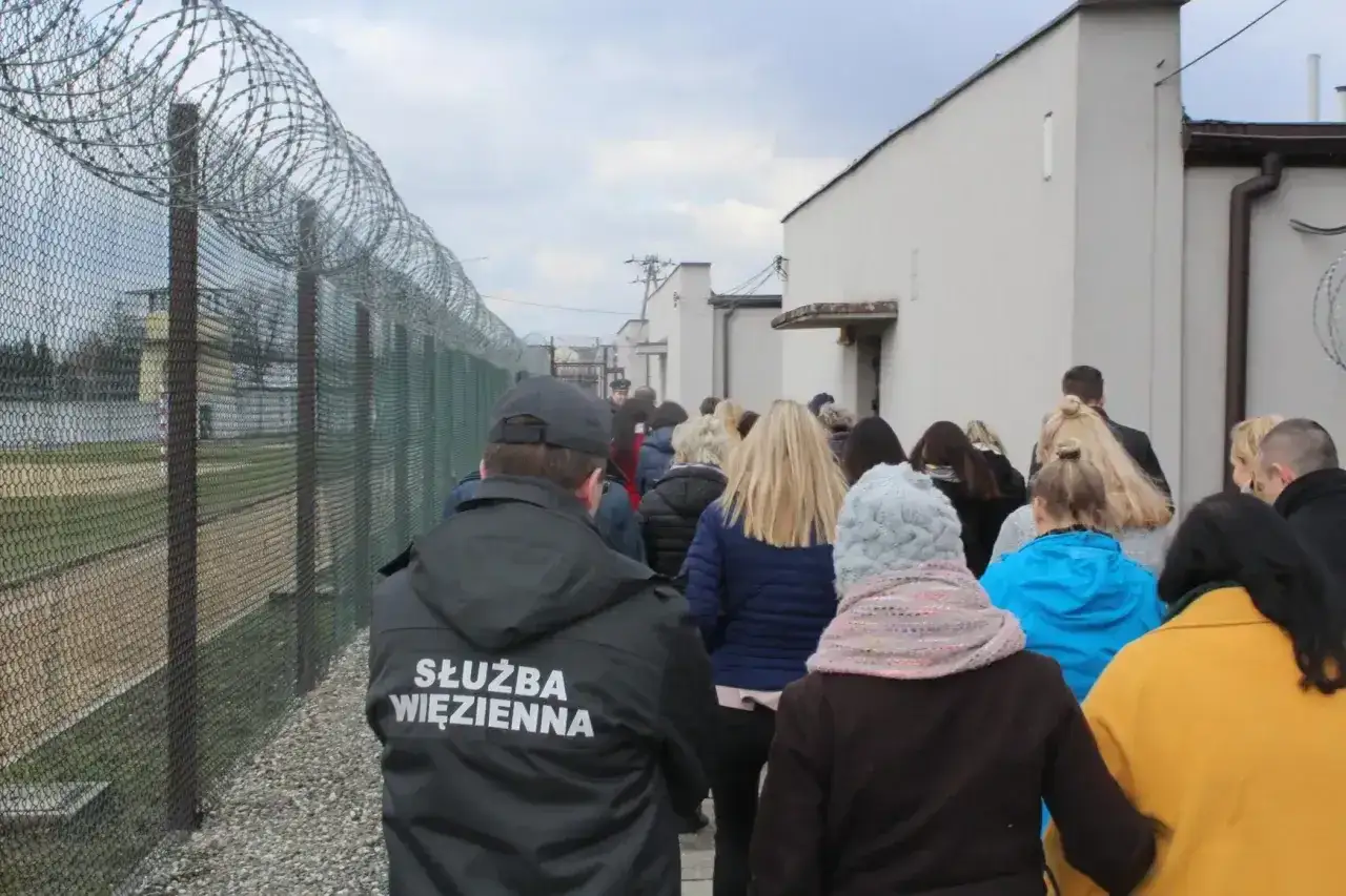Grupa osób wchodzi na teren więzienia. Strażnik w kurtce z napisem 