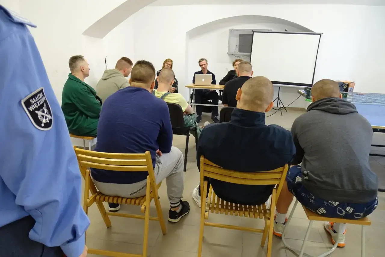 Grupa mężczyzn, w tym więźniowie i funkcjonariusz Służby Więziennej, słucha wykładu. Na ekranie projekcyjnym widnieje napis 