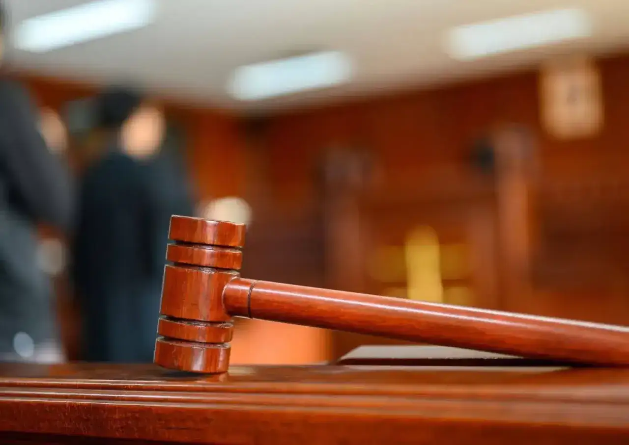 Młotek sędziowski na drewnianym stole, symbol sprawiedliwości i dochodzenia odszkodowania. W tle niewyraźne sylwetki ludzi.